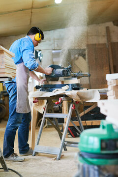 Caucasian young adult man operating circular saw in woodworking workshop, wearing protective ear muffs and apron, cutting wooden planks with sawdust flying in air, focused on task