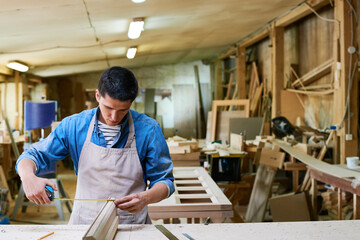 Young adult Caucasian man measuring wooden plank with tape measure in carpentry workshop, focusing on precise measurement while working on woodworking project surrounded by tools and materials