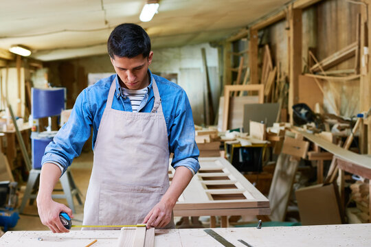 Young adult Caucasian man measuring wooden plank with tape measure in carpentry workshop, focusing on precise measurement while working on woodworking project surrounded by tools and materials