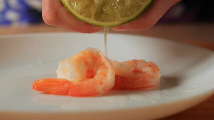 hand squeezing a lime half. A single drop of juice is captured falling onto a plate of cooked shrimp - Powered by Adobe