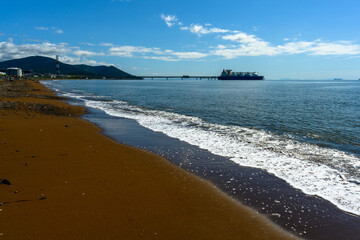Sunny shoreline with gentle waves and a distant ship near hills under a clear blue sky