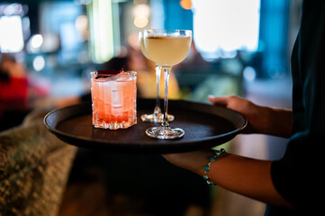 A person's hands holding a black round tray with two different cocktails on it, with a bar interior in the soft-focus background.