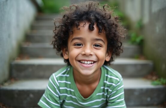 Cute smiling boy with curly hair sits on outdoor steps. Happy child enjoys fun time outside. Kid has good mood, plays in warm weather. Young person has joyful expression. Mixed race kid face.