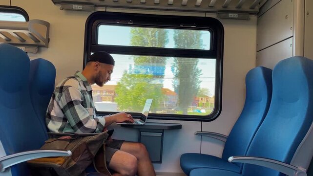 Young man with backpack sitting in train using laptop, working while passing through suburban landscape. Digital nomad concept. 4k horizontal footage