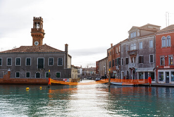 Scenic view of Murano Island with historic buildings and a clock tower reflected in the tranquil water, showcasing the charm of Venetian architecture and vibrant colors