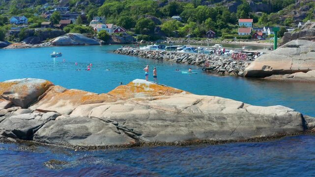 People swimming in summertime in Norway in Fjord. Sandefjord nature aerial video over rocky coastline and small boat harbour. Kids standing on a cliff ready to jump in