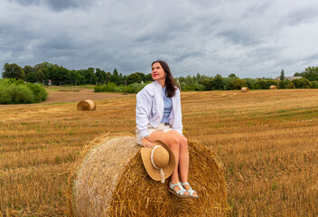 Young woman in casual clothes and summer sandals sits on a hay bale holding her straw hat. The...