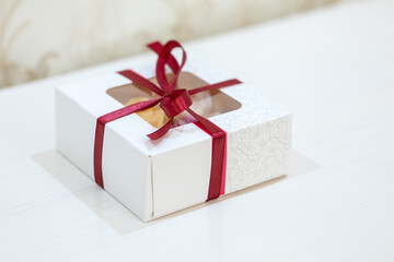 Small white cardboard gift box tied with a red satin ribbon, featuring a clear window and decorative pattern on a light surface