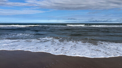 Image of a beach with a horizon separating the sea from the sky. Waves break on the shore, creating white foam that spreads across the wet sand, and the sky is partially light blue with clouds.