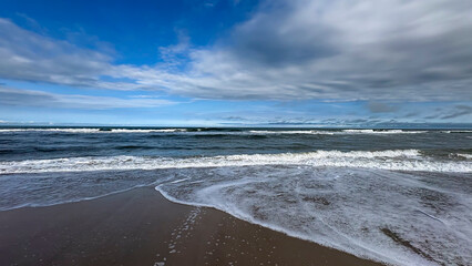 Image of a beach with a horizon separating the sea from the sky. Waves break on the shore, creating white foam that spreads across the wet sand, and the sky is partially light blue with clouds.