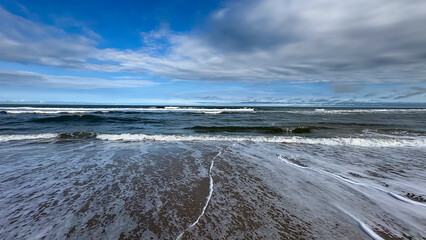 Image of a beach with a horizon separating the sea from the sky. Waves break on the shore, creating white foam that spreads across the wet sand, and the sky is partially light blue with clouds.