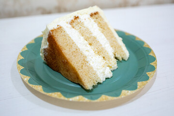Single slice of layered vanilla sponge cake with rich white cream frosting on a teal and gold decorative paper plate