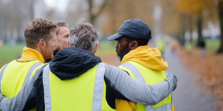 Group of diverse men wearing high visibility vests embracing outdoors in autumn park setting with blurred background - Powered by Adobe