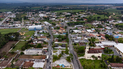 A drone shot of the town of Balsa Nova, near Curitiba, on a cloudy day, shows a town square, a church, houses and commercial buildings, and the view extends to the hills on the horizon.