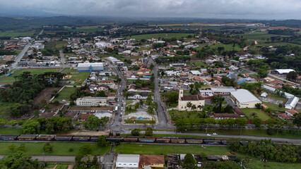 A drone shot of the town of Balsa Nova, near Curitiba, on a cloudy day, shows a town square, a church, houses and commercial buildings, and the view extends to the hills on the horizon.