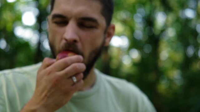 Young man in light green t shirt sitting on bench in park, eating red apple surrounded by green trees and sunlight. 4k horizontal footage