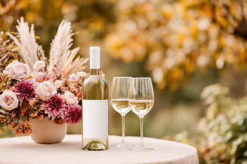 Two glasses of white wine and a bottle with blank mockup label on a floral-decorated table, set against a fall outdoor backdrop