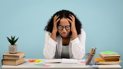 Tired African American female student cries over her books and notebooks while preparing for a challenging college exam. She sits at a desk surrounded by study materials and office supplies.