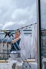 Young woman hanging laundry on balcony drying rack and clothespins, air-drying for energy saving and eco-friendly living, casual apartment lifestyle with city and mountain view, candid domestic scene