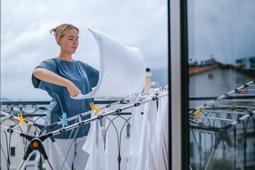 Young woman hanging laundry on balcony drying rack and clothespins, air-drying for energy saving and eco-friendly living, casual apartment lifestyle with city and mountain view, candid domestic scene