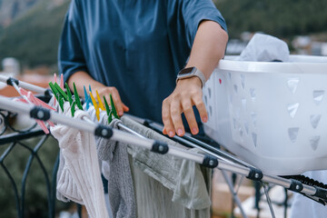 Young woman hanging laundry on balcony drying rack and clothespins, air-drying for energy saving and eco-friendly living, casual apartment lifestyle with city and mountain view, candid domestic scene