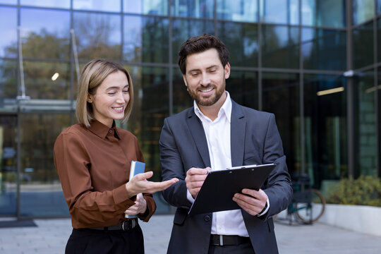 Two smiling business professionals collaborating on a project, reviewing documents on a clipboard outside a modern glass office, showcasing teamwork and partnership in a corporate setting