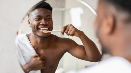 African American man stands near a mirror in a stylish bathroom, happily brushing his teeth with a toothbrush. He is wrapped in a towel, embracing his morning routine with joy.
