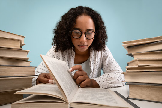 A focused African American female student sits at a desk surrounded by stacks of books. She is deeply engaged in reading and studying for an upcoming exam against a blue background.