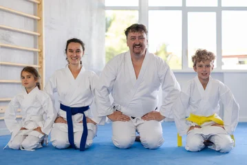 Selbstklebende Fototapeten Kampfkunst Smiling parents with preteen children, son and daughter, wearing traditional white kimonos sitting on knees on tatami in gym, ready for martial arts training. Sports family concept  © JackF