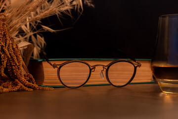 Stylish reading glasses resting on a vintage book, with a glass of whisky partially visible on the right. A cozy and intellectual still life scene, suggesting relaxation or evening study