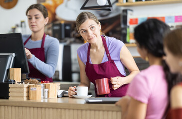 Polite young barista pouring coffee into cup while clients waiting at counter in cafeteria