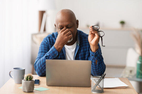 Mature African American man is sitting at a laptop, rubbing his nosebridge while holding glasses. He appears tired after working on the computer in a modern indoor office environment.