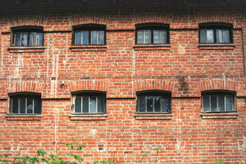 Old brick building facade with small framed windows arranged in two rows. Weathered red brick wall with aged texture. Vntage architecture