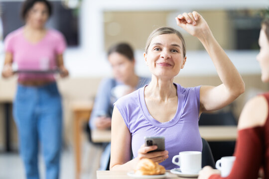 Positive young woman calling for waitress while sitting at table in bright cafeteria