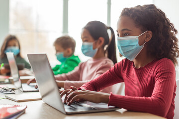 In a contemporary classroom, an African American schoolgirl focuses on her laptop while participating in online learning. Diverse students, all wearing face masks, engage at their desks.