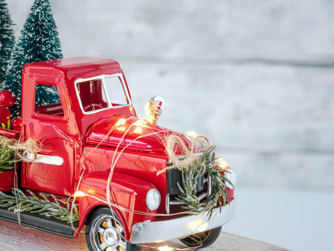 Background for personalized Christmas card. Red country toy truck and twinkle fairy lights with trees and festive decorations on white backdrop. Vintage sleigh bells, greenery wreath, and red berries.