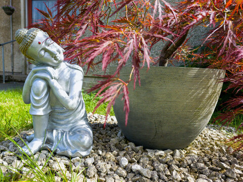 Ceramic Buddha in garden in front of Japanese maple tree with red leaves in large gray pot. Siddhartha Gautama, Buddha&thinsp;the awakened one. Wandering ascetic, religious teacher,  Asia founded Buddhism.