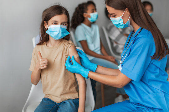 In a clinic setting, a medical worker administers a coronavirus vaccine to a preteen girl. She smiles and gives a thumbs up gesture while wearing a mask. Other children wait nearby.