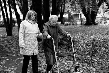 Woman and her elderly mother with walker taking a walk in the park covered in autumn leaves, spending family time together