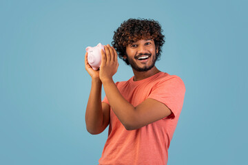 A cheerful young indian man with curly hair holds a piggy bank next to his ear, showcasing joy and...