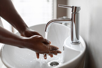 An unrecognizable African man cleans his hands under running water in a contemporary bathroom. The focus is on his arms and the efficient handwashing process taking place.