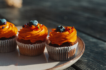 Delicious cupcakes with orange frosting and blueberry toppings on a plate on a rustic dark wooden table. These tasty sweets and treats are perfect for a special occasion like Halloween holiday.
