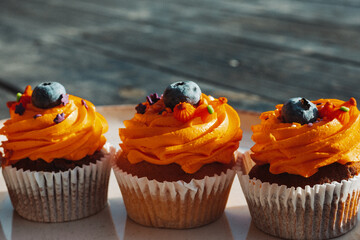 Delicious cupcakes with orange frosting and blueberry toppings on a plate on a rustic dark wooden table. Tasty sweets, treats, perfect for Halloween.
