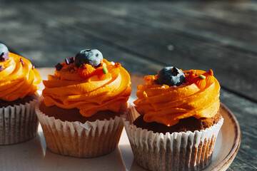Delicious cupcakes with orange frosting and blueberry toppings on a plate on a rustic dark wooden table. Tasty sweets, treats, perfect for Halloween.