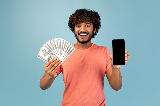 A young Indian man in a casual pink shirt smiles broadly while holding a blank smartphone screen in one hand and cash in the other against a blue studio background.