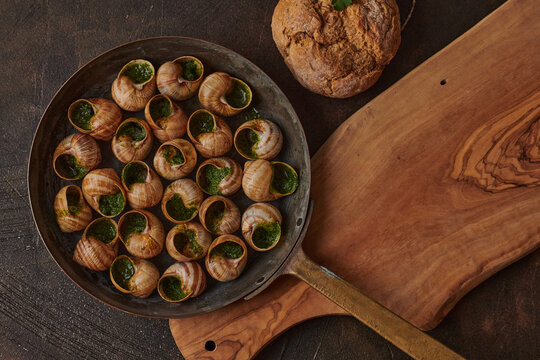 Gourmet dish featuring escargot in a rustic pan, garnished with herb butter, served alongside freshly baked bread on wooden board
