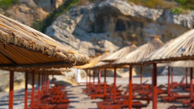 Sandy beach with straw umbrellas focusing from rocky background. Idyllic empty beach with rows of red sun loungers and straw umbrellas on a sunny day