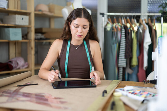 Fashion designer concentrates on drawing a model of clothes and patterns on a tablet. Process of creating an individual collection of clothes in a sewing workshop - Powered by Adobe
