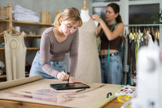 Thoughtful young female dressmaker working on digital sketch of new dress using tablet in fashion studio while colleague working on fitting dress on mannequin in background