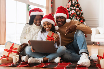 A joyful black family sits on the floor, wearing red Santa hats while making a video call on a laptop. They celebrate Christmas together, surrounded by gifts and a festive atmosphere.
